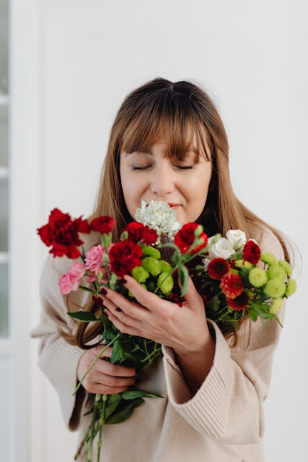 A woman with light brown hair and bangs, dressed in a beige coat, is holding a vibrant bouquet of fresh flowers close to her face, with her eyes closed and a gentle smile. The bouquet includes red car