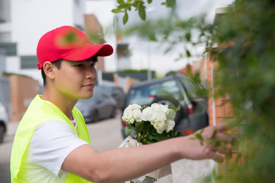 A young delivery person wearing a red cap and high-visibility yellow vest is standing outdoors on a residential street, holding a bouquet of fresh white hydrangeas wrapped in white paper. The arrangem