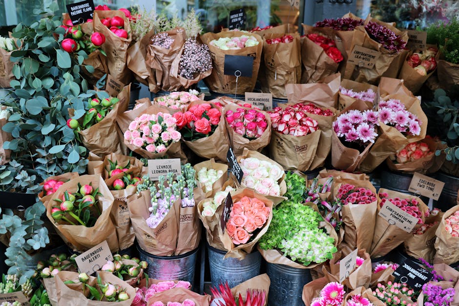 A vibrant display of fresh flower bouquets arranged in rustic brown paper wraps and placed in metal buckets, featuring a variety of blooms in pink, red, white, and purple shades. The flowers include r