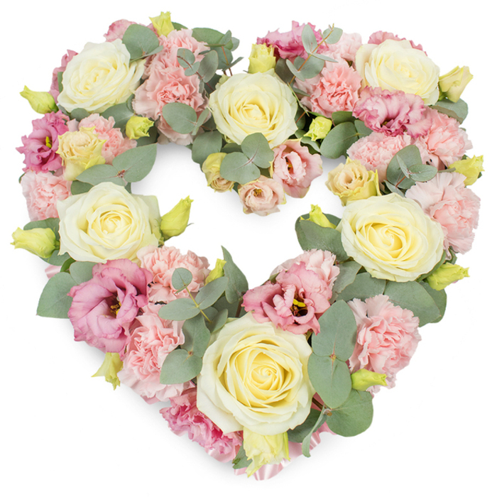 This photograph shows a luxuriously full, heart-shaped wreath, created as a tribute piece with the refined touch one might expect from a dedicated Radlett florist. The heart is perfectly proportioned, its outline entirely softened by an abundance of flowers worked closely together so no underlying structure is visible. Cream and buttery pale yellow roses provide the main focal points, their velvety petals arranged in tight spirals that open gradually, giving a sense of calm and measured elegance. Around and between these roses, clusters of pastel pink carnations are carefully positioned, their delicately frilled petals creating a soft, layered texture reminiscent of gently folded fabric. To deepen the colour palette, lisianthus in rich pink and soft mauve tones are interspersed throughout, their open, cup-like blossoms adding both volume and visual rhythm, as if echoing the gentle ebb and flow of conversation at a remembrance service. Silvery eucalyptus foliage winds throughout the design, its round, slightly dusty green leaves bringing cool contrast and a subtle, natural structure, much like the peaceful greenery surrounding Radlett's residential streets. Small sprays of yellow-green buds appear at intervals, suggesting freshness and the continuing cycles of nature. The overall impression is serene and contemplative, with pastel shades of pink, cream, and green merging into a cohesive, soothing whole. This heart-shaped wreath would feel very much at home at a Radlett funeral, graveside ceremony, or memorial in nearby villages, a floral expression chosen to honour a special relationship and to surround a final farewell with gentle beauty and quiet love.